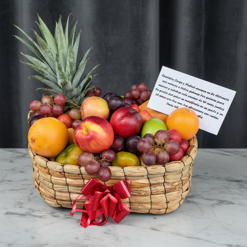 Canasta tejida llena de frutas frescas como piña, naranjas, manzanas, nectarinas, uvas y limones, decorada con lazo rojo y tarjeta de dedicatoria sobre mesa de mármol con fondo gris