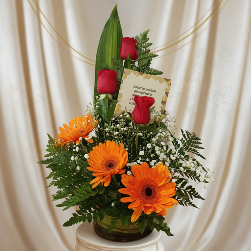 Arreglo floral en jarrón de vidrio con gerberas naranjas, rosas rojas, follaje verde y tarjeta sobre fondo de cortina beige