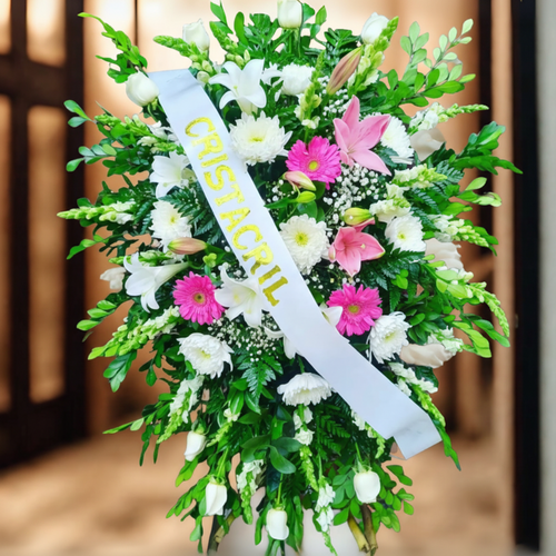 Arreglo floral fúnebre grande con flores blancas, rosas y fucsias, follaje verde abundante y cinta blanca diagonal con texto dorado