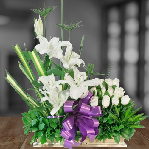Arreglo floral rectangular con lirios y rosas blancas, follaje verde y gran lazo morado sobre mesa de madera, fondo interior desenfocado