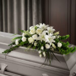 Centro de flores fúnebres con lirios y rosas blancas sobre un ataúd gris en una sala con cortinas oscuras