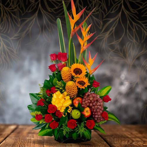 Arreglo floral vertical con heliconias, girasoles, rosas rojas, piñas decorativas, uvas y manzanas sobre mesa de madera con fondo oscuro