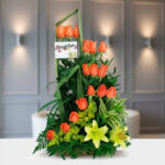 Arreglo floral vertical con rosas naranjas, lirios amarillos y follaje verde sobre mesa blanca en interior elegante
