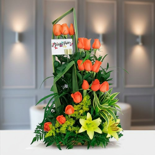 Arreglo floral vertical con rosas naranjas, lirios amarillos y follaje verde sobre mesa blanca en interior elegante