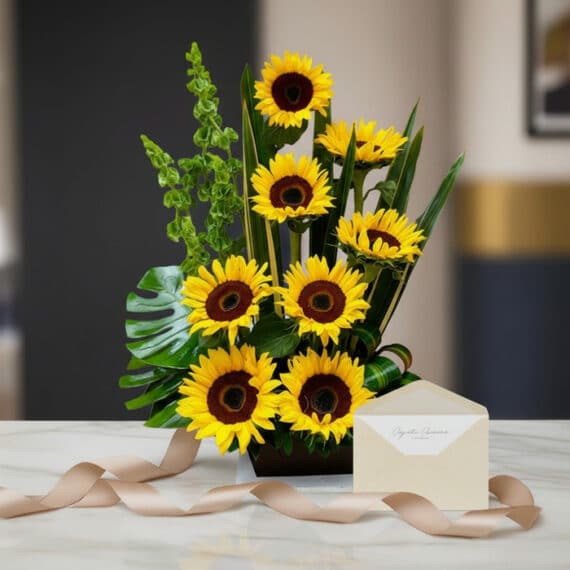 Arreglo floral de girasoles Bengala vibrantes con follaje verde exótico, presentado en un jarrón oscuro sobre una mesa de már