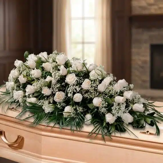 Elegante Arreglo Fúnebre Cubre Caja Cielo Rosas Blancas Arreglo fúnebre cubre caja con rosas blancas y follaje verde, sobre un ataúd de madera clara en una sala de velación premium