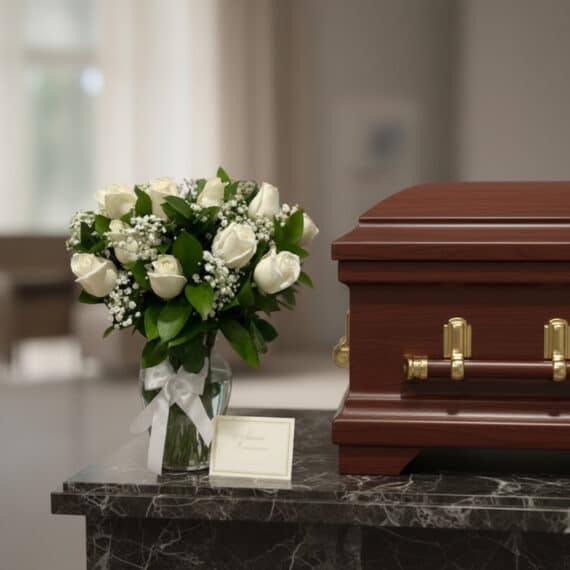 Arreglo floral fúnebre 'Pétalos del Alma' con rosas blancas y follaje, junto a un ataúd de madera en una sala de velación pre