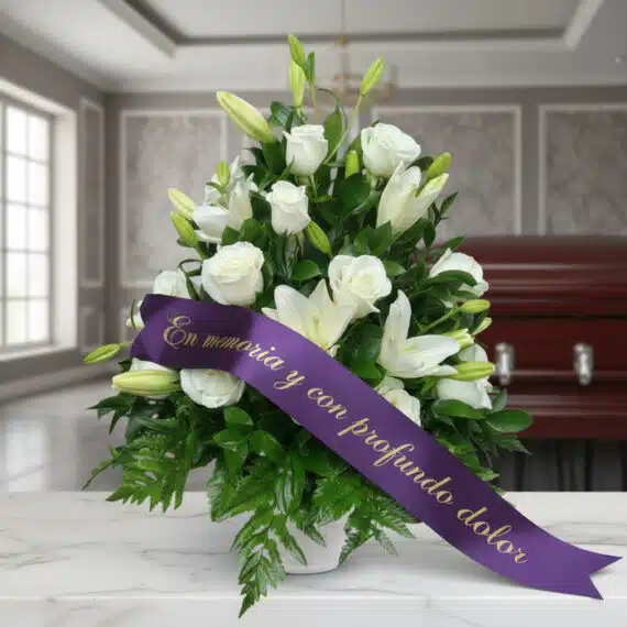 Arreglo fúnebre Piedad con rosas y lirios blancos, cinta morada con mensaje de condolencia, en sala de velación premium.