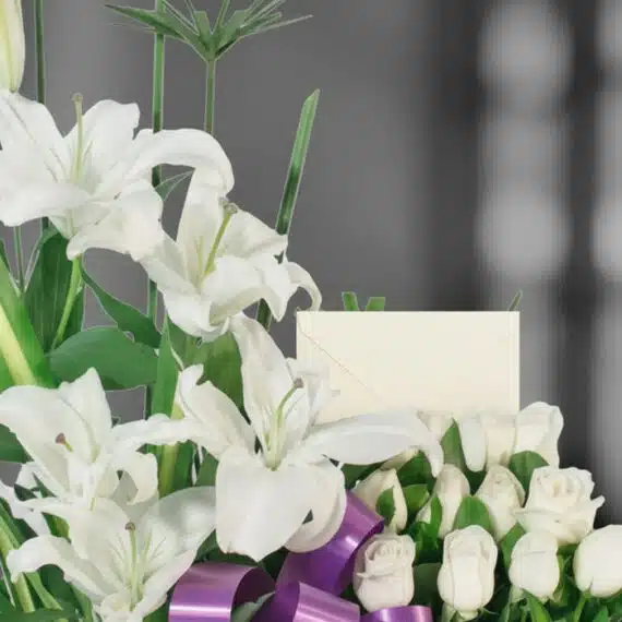 Primer plano de Arreglo Fúnebre Pureza con lirios blancos, rosas y cinta morada, rocío sutil en pétalos y follaje, profundida