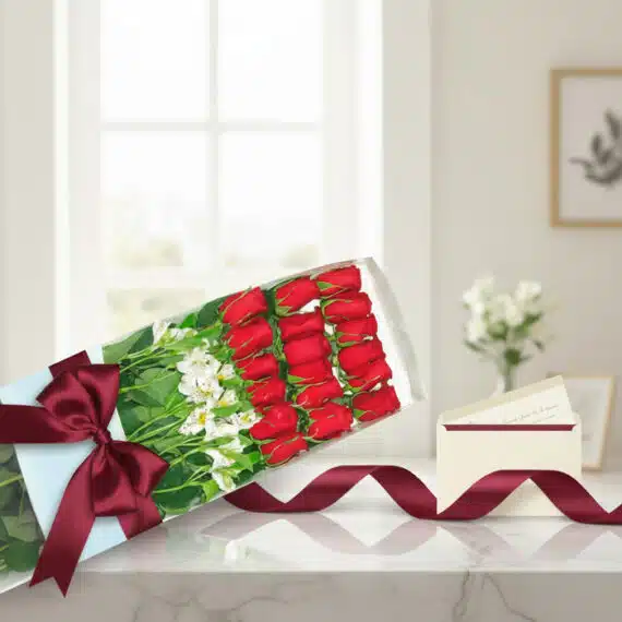 Caja de 18 rosas rojas frescas con alstroemerias blancas, lazo de satén burdeos y tarjeta de regalo en sobre, sobre mármol.