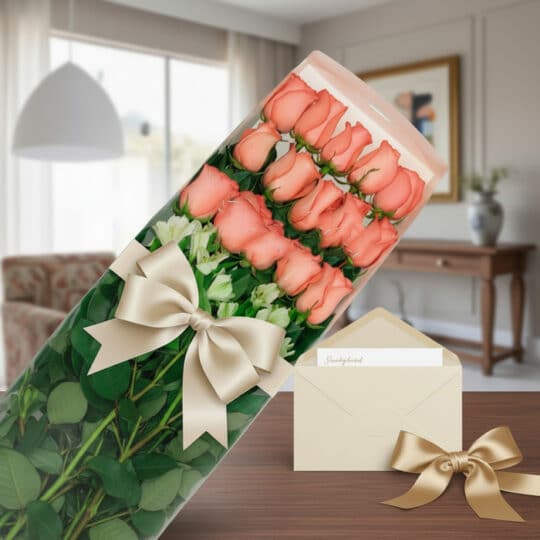 Caja de 18 rosas rosadas frescas con follaje verde, lazo de satén beige y tarjeta de regalo en un sobre, sobre una mesa de ma