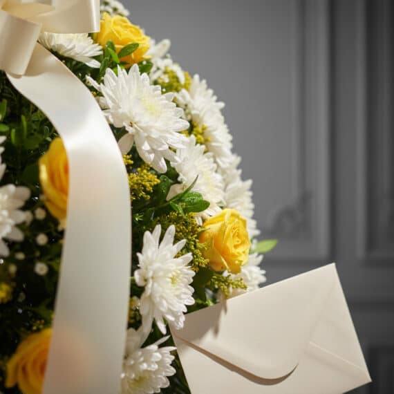 Primer plano de una corona fúnebre con rosas amarillas, crisantemos blancos y follaje verde, con una cinta satinada y un sobr
