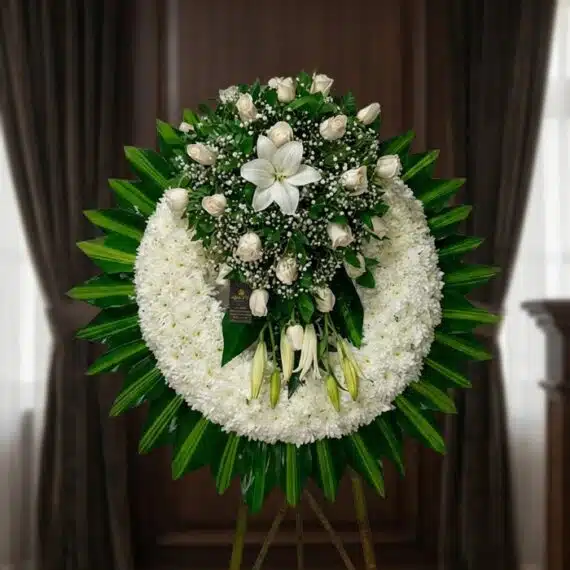 Elegante Corona Fúnebre con Rosas y Lirios Blancos para Homenajes Corona fúnebre grande con rosas blancas, lirios y follaje verde, presentada en una sala de velación premium con luz natural s