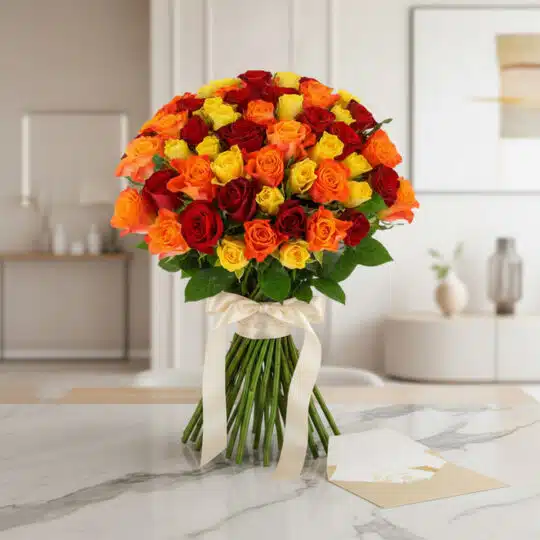 Ramo SYLVANA con 60 rosas rojas, naranjas y amarillas, atado con cinta de satén crema, sobre mesa de mármol con tarjeta de re